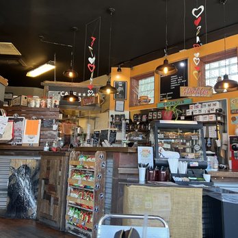 Cafe Urbano - Order counter - The image shows the interior of a cafe with a wooden counter, menu boards on the wall, and a display case filled with baked goods. Decorative hanging hearts are suspended from the ceiling, and there are various coffee-making equipment and supplies visible. The space has warm lighting, a window allowing natural light, and a cozy, inviting atmosphere. - Cafes Near Me - Chicago, Illinois