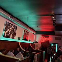 Inside - A dimly lit indoor space featuring artwork on the walls, neon signage, and groups of people seated and standing, suggesting a bar or restaurant setting.