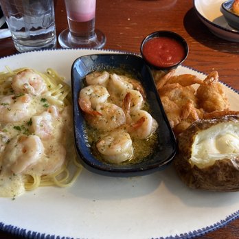 Red Lobster - Seaside Shrimp Trio - A seafood meal includes shrimp in a creamy white sauce served over pasta, fried shrimp with a side of cocktail sauce, and a baked potato topped with butter. The dish is presented on a white oval plate on a wooden table, with drinks and additional dishes visible in the background. - Seafood Near Me - Albuquerque, New Mexico