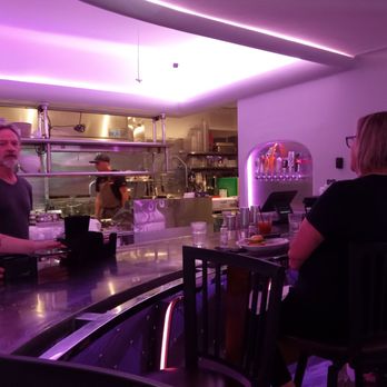 Bisbee Royale - The counter from my seat - The image shows a bar interior with patrons seated at the counter, including a woman with glasses and a man with gray hair. Behind the bar, a chef is working in the kitchen area, which is partially visible. The ceiling and walls are lit with purple lighting, creating a modern atmosphere. The bar counter has glasses, plates, and drinks, and the background features shelves with bottles and bar equipment. - Music Venues Near Me - Bisbee, Arizona