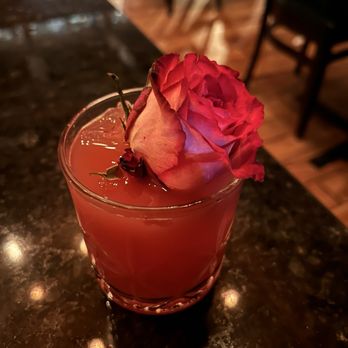 Prohibition - Bourbon & The Beast - A pinkish cocktail in a short glass features a dried rose as a garnish. The drink is on a dark, polished bar counter with warm lighting, and blurred chairs and tables are visible in the background, indicating a bar or restaurant environment. - Cocktail Bars Near Me - Murray, Utah