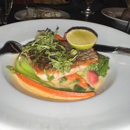 Sea Bass - A plated dish displays a piece of grilled salmon with crispy skin, placed on a bed of cooked vegetables including carrots, celery, and greens. The dish is garnished with microgreens and a lime wedge on top. The presentation is on a white, round plate, with a dark table setting and other dishes and glasses visible in the background.