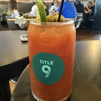 Title 9 - A vibrant red cocktail with garnishes, including a cucumber slice, green olives, and a star-shaped skewer, on a table in a lively dining area. - Sports Bars Near Me - Pittsburgh, Pennsylvania