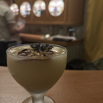 Daydream - A cocktail served in a stemmed glass with a creamy yellow hue, topped with a garnish of lavender sprigs and star anise, placed on a wooden table. The background shows a blurred restaurant interior with a person and decorative circular mirrors on a wooden wall. - Cocktail Bars Near Me - Albuquerque, New Mexico