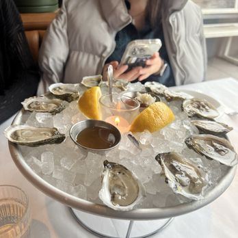The Snail - oysters - A woman is seated at a table with a large seafood platter featuring oysters on ice, lemon wedges, and dipping sauces. She is holding a smartphone and appears to be engaged with it. The setting includes a white tablecloth, glassware, and a window in the background, suggesting a restaurant environment. - American Near Me - Brooklyn, New York