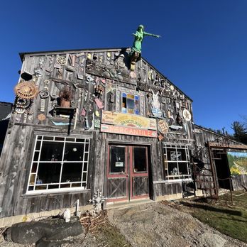 Three Sisters Sanctuary - Three Sisters Sanctuary - A weathered wooden building decorated with metal art and sculptures, featuring a sign for a sanctuary and mercantile, with a figure of a girl on the roof. - Parks Near Me - Goshen, Massachusetts
