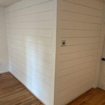 Knotty's Custom Carpentry - Final product!  Real shiplap, NOT tongue and groove flooring! - The image shows a corner of an interior room with white shiplap wall paneling installed on two adjacent walls. The paneling consists of horizontal wooden planks with visible lines. The flooring is hardwood with a natural wood finish. An electrical outlet is present on one wall, and a door is partially visible on the right side of the image. The ceiling appears to be sloped, indicating an attic or upper floor space. - Handyman Near Me