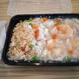 Egg Rolls - A black plastic takeout container contains shrimp with lobster sauce, rice, and mixed vegetables, placed on a wooden table with a napkin on the side.