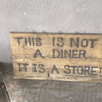 Maggie's Diner - This says it all - A rustic wooden sign with black lettering reads, 'This is not a diner, it is a store!' placed on a concrete surface near a wall. - Souvenir Shops Near Me - Madrid, New Mexico