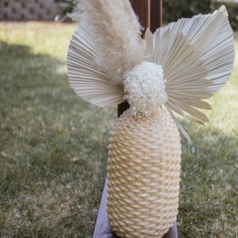 Light and Well - A decorative floral arrangement includes dried pampas grass, paper fan-like leaves, and a white pom-pom, all placed in a woven vase. The arrangement is positioned on a wooden base outdoors, with grass and trees visible in the background, suggesting it is part of an outdoor event or display. - Floral Designers Near Me
