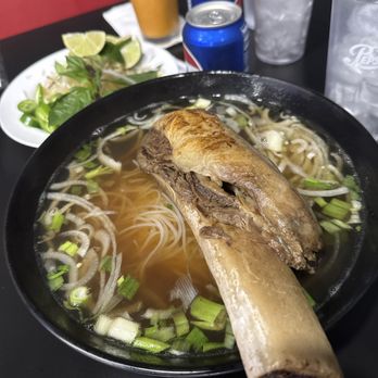 84 Viet - Dinosaur Pho - A bowl of Vietnamese pho contains rice noodles, chopped green onions, and a large beef bone placed on top. The broth is clear, and the ingredients are visible within the bowl. In the background, there are cans of soda, glasses of ice water, a glass of orange-colored drink, and a plate with lime wedges and greens. The setting appears to be a casual dining environment, likely at a Vietnamese restaurant. - Vietnamese Near Me - Berkeley, California