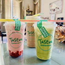 32oz House Special - Three beverages in clear plastic cups with straws are positioned on a reflective table inside a room. The drinks are pink with red syrup at the bottom, beige, and green, each with a green Tamper Evident label. The room has curtains with valances, a window, and decorative items on shelves, indicating an indoor setting such as a home or cafe.