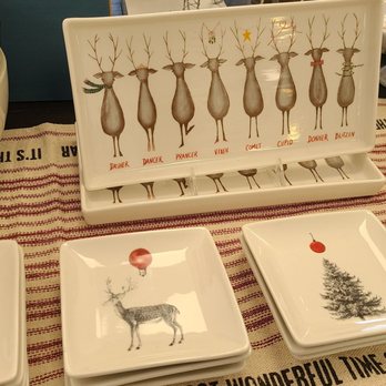 Sweet Things Chocolates - So cute! - The image shows several ceramic plates with holiday motifs, including a reindeer, Christmas tree, and ornaments, placed on a striped tablecloth. A decorative card with reindeer illustrations labeled with names is positioned behind the plates. To the left, there are stacked white bowls with a partial message visible, and to the right, white mugs with red interiors are arranged. The setting appears to be a display of holiday tableware. - Candy Stores Near Me - Indianapolis, Indiana