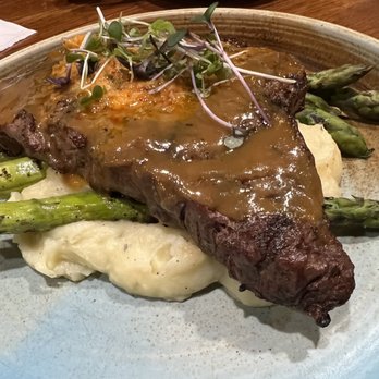 Subourbon - Rib-eye - A dish with a cooked rib-eye steak topped with gravy, placed on a bed of mashed potatoes. The plate also includes roasted asparagus spears and a garnish of microgreens on top of the steak. The presentation is on a ceramic plate, with a wooden table surface visible in the background. - Southern Near Me - Columbus, Ohio