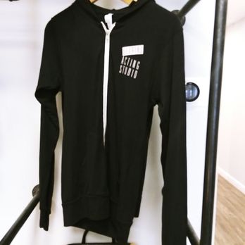 Dearing Acting Studio - A black zip-up hoodie with a white zipper and white text on the chest is displayed hanging on a wooden hanger. The hoodie is positioned on a metal clothing rack in an indoor setting, with a basket visible below and a white wall in the background. - Performing Arts Near Me - Phoenix, Arizona