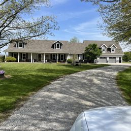 A large, two-story house with a front porch and attached garage is situated in a spacious yard with green grass and mature trees. The gravel driveway curves toward the garage, and the sky is clear with some clouds. The yard includes a flower pot and benches, and a small shed is visible in the background.