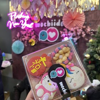 Mochiido - A hand holds a clear plastic box containing decorated cookies, including a Hello Kitty cookie, a cookie with yellow icing and symbols, and colorful, bead-like treats. The background features a decorated wall with pink, gold, and pastel paper decorations, a neon sign reading 'Taking Care of You,' and the Mochiido logo, indicating a celebration or event at the Asian fusion bakery in New York. - Donuts Near Me - Flushing, New York