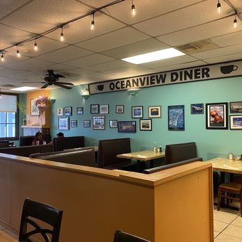 Oceanview Diner - Inside interior after closing - The photo depicts the interior of Oceanview Diner, featuring a light blue wall decorated with framed pictures and posters. String lights hang from the ceiling, and there are booth seats with dark upholstery along the wall. A window with blinds is visible on the left, and a small corner with balloons and a fireplace is in the background. The ceiling has a ceiling fan and fluorescent lighting. - Breakfast & Brunch Near Me - Norfolk, Virginia