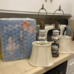 Shipped to Cali - Vintage lamps with floral designs, a boxed doll wrapped in bubble wrap, and kitchen appliances are arranged on a kitchen countertop.