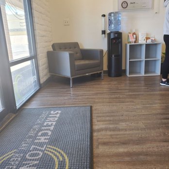Stretch Zone - The image shows a waiting area with a gray upholstered armchair positioned against a white wall. Next to the chair is a black water cooler with a large water bottle on top. To the right of the cooler, there is a white shelf with informational pamphlets and small items. The area is illuminated by natural light coming through a glass door on the left, which has a mat with the logo and name of the business, 'Stretch Zone.' The floor is wood laminate, and the wall behind the water cooler has a framed certificate. A person is partially visible on the right side of the image. - Trainers Near Me - Centennial, Colorado