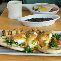 A breakfast meal includes eggs Benedict topped with hollandaise sauce and garnished with greens, served with roasted potatoes. Additional dishes include a bowl of black beans and a dish with fried eggs and meat. The meal is set on a wooden table with a white mug and utensils visible.
