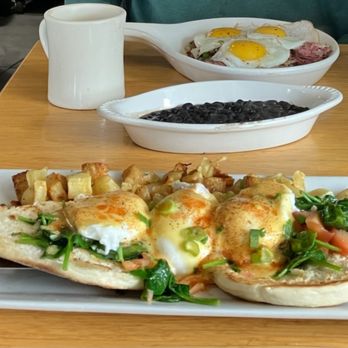 Hangar 12 - A breakfast meal includes eggs Benedict topped with hollandaise sauce and garnished with greens, served with roasted potatoes. Additional dishes include a bowl of black beans and a dish with fried eggs and meat. The meal is set on a wooden table with a white mug and utensils visible. - Breakfast & Brunch Near Me - Mansfield, Massachusetts