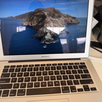 iFixOnWheelz - MacBook - A MacBook Air laptop is positioned on a desk, with its screen displaying a landscape wallpaper of a mountain surrounded by ocean. The keyboard is visible, and the background includes various office or workspace items, indicating an indoor setting. - Mobile Phone Repair Near Me - Cambridge, Massachusetts