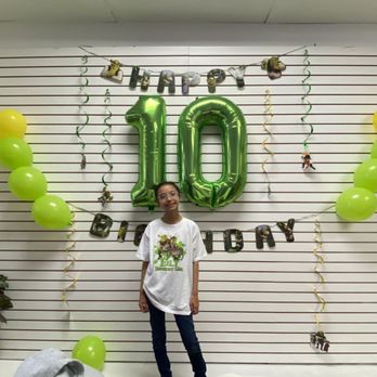 Slime World - A girl poses in front of a birthday backdrop decorated with balloons, a large green '10' balloon, and hanging banners that say 'Happy Birthday.' - Party & Event Planning Near Me - Fairfield, California