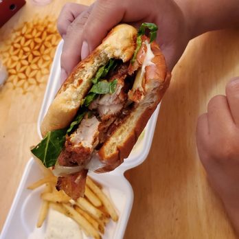 Birdcall - All Natural Chicken - Deluxe chicken sandwich - A chicken sandwich with lettuce, bacon, and sauce is held in a person's hand over a takeout container with fries and dipping sauce. - Chicken Shop Near Me - Denver, Colorado