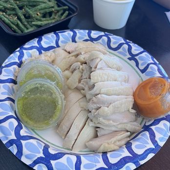 Cluck2Go - The image features a plate of sliced poached chicken served with two small containers of green sauces, a cup of clear broth, and a side of cooked green beans. The plate has a blue and white patterned border, and the setting appears to be a casual dining restaurant. Additional food items and a person's arm are visible in the background. - Singaporean Near Me - Pasadena, California