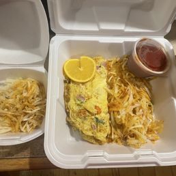 Breakfast Burrito - A takeout meal served in a white foam container includes an omelette garnished with lemon slices, hash browns, and a small cup of red sauce. An additional portion of hash browns is in a separate foam container, and a disposable coffee cup with a lid is placed nearby. The meal appears to be breakfast from a restaurant or cafe.