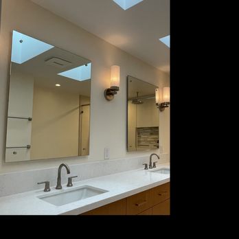 PCD Remodeling - His and hers sinks, quartz countertops, floating mirrors, custom cabinetry - The image shows a bathroom with a long white countertop containing two sinks, each with a brushed nickel faucet. Above the sinks are two rectangular mirrors, flanked by wall-mounted light fixtures. The ceiling has multiple skylights allowing natural light into the space. The background includes a shower area with a glass door and a tiled wall. The vanity has wooden cabinetry below the countertop. - General Contractors Near Me - La Jolla, California