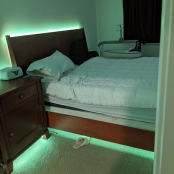 Abc Moving & Contracting - The image shows a bedroom with a bed that has white bedding and a pillow, positioned against a wall with green LED lighting behind the headboard. A wooden nightstand is beside the bed, holding a white device. A black backpack rests on the bed, and a pair of white slippers is on the carpeted floor. Near the window, which is covered by dark curtains, there is a baby bassinet with a small lamp attached. The room has a calm, dimly lit atmosphere with a focus on the bed and surrounding furniture. - Office Cleaning Near Me