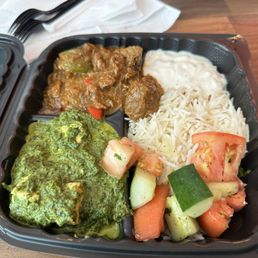 Lamb and spinach - A black plastic takeout container contains a serving of white rice, a portion of curry with vegetables, cooked spinach with a green sauce, and a vegetable salad with chopped tomatoes, cucumbers, and carrots. The container is placed on a wooden surface, with white napkins and utensils visible in the background.
