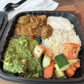 SpiceX - Lamb and spinach - A black plastic takeout container contains a serving of white rice, a portion of curry with vegetables, cooked spinach with a green sauce, and a vegetable salad with chopped tomatoes, cucumbers, and carrots. The container is placed on a wooden surface, with white napkins and utensils visible in the background. - Indian Near Me - Washington, DC, District of Columbia