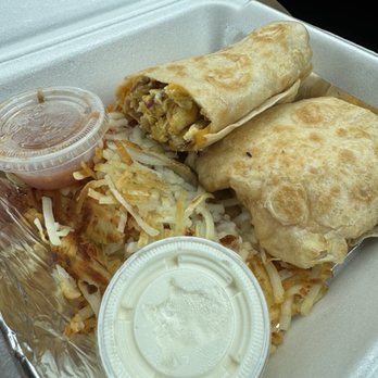 Lina's cafe - The image shows a foam takeout container with a breakfast meal. Inside, there is a partially eaten breakfast burrito with visible scrambled eggs, meat, and vegetables. Alongside, there are crispy hash browns topped with shredded cheese. Two small plastic cups of dipping sauces, one pink and one white, are also present. The meal appears to be served for takeout, with the food arranged on a bed of foil. - New American Near Me - Slinger, Wisconsin