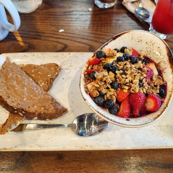First Watch - A.M. superfood bowl. - A breakfast plate includes a cooked chicken leg with gravy, a scoop of mashed sweet potato, and a bowl of yogurt topped with strawberries, blueberries, and granola. The meal is presented on a rectangular white ceramic plate placed on a wooden table, with drinks and utensils visible in the background. - Breakfast & Brunch Near Me - Tucson, Arizona