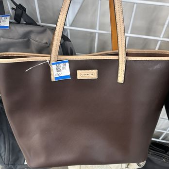 Savers - A brown Coach tote bag with beige leather handles is hanging on a metal grid display rack in a retail store. The bag features a small Coach logo patch on the front and has a price tag showing $66.99. The background includes other bags and store fixtures, indicating a retail environment for handbags and accessories. - Thrift Stores Near Me - Santa Clara, California