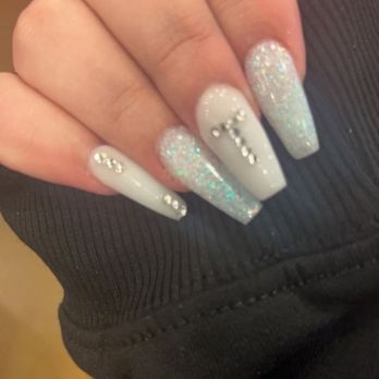 She Shed - The image displays a close-up of a hand with long, manicured nails painted in white and glittery shades. Some nails are decorated with rhinestones arranged in patterns. The hand is partially covered by a black, ribbed fabric, possibly a sleeve. The background is blurred and appears to be indoors with warm lighting. - Nail Salons Near Me - Albuquerque, New Mexico