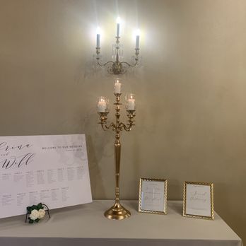 AGS Event Creations - A wedding seating chart display features a large white board with the names 'Sabrina and Will' and a seating arrangement list. The table also holds a tall, gold-colored candelabrum with multiple lit candles, and two small framed signs. The setup is against a plain wall, with a white tablecloth covering the table. - Wedding Planning Near Me - Mundelein, Illinois