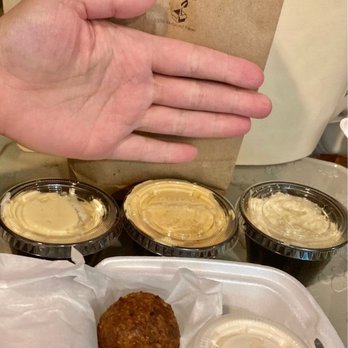 Cafe Baladi - Cafe Baladi -- app sampler - The image shows a hand holding a paper bag from Cafe Baladi, with three small containers of hummus placed on a table in front. The containers are filled with hummus, and a white tray contains two fried falafel balls wrapped in paper. The background includes a beige wall and part of a white object, possibly a bag or cushion. - Lebanese Near Me - Malvern, Pennsylvania