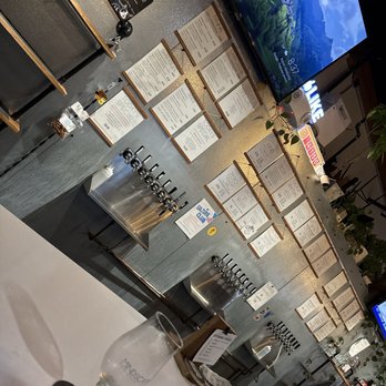 Mindscape Fermentations - The image shows a bar area with multiple stainless steel beer taps mounted on a counter. Behind the taps, there are numerous menu boards attached to the wall, listing various drink options, specials, and prices. A large television screen is mounted on the wall, displaying a scenic landscape. The bar has some decorative elements, including small plants and signage, and a glass with a straw is visible in the foreground. - Breweries Near Me - Rocklin, California