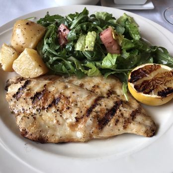 Athos - I ordered "WHOLE" bronzino and asked them to filet it - A white plate holds a grilled fish fillet with visible grill marks, accompanied by a halved lemon with charred surface, boiled potatoes, and a mixed green salad with arugula, lettuce, and bacon bits. The dish is presented on a white tablecloth with a wine glass nearby. - Greek Near Me - Albany, New York