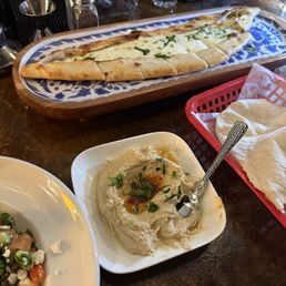Hummus, cheese pide - The image shows a restaurant table with a plate of hummus garnished with herbs and olive oil, a cheese pide on a decorative tray, and a side dish with vegetables.