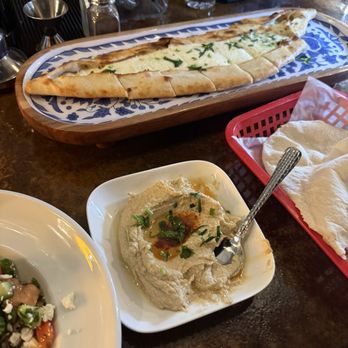 Selda - Hummus, cheese pide - The image shows a restaurant table with a plate of hummus garnished with herbs and olive oil, a cheese pide on a decorative tray, and a side dish with vegetables. - Mediterranean Near Me - Irving, Texas