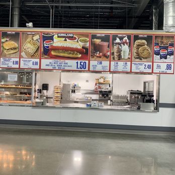 Costco - Lots of space - A Costco food court area features a stainless steel serving counter with various equipment and supplies. Above the counter, menu boards display food items such as hot dogs, sandwiches, salads, ice cream, and beverages, along with prices and calorie information. The ceiling has exposed industrial piping and ductwork, and the floor is polished concrete. The setup appears clean and organized, typical of a Costco food court. - Wholesale Stores Near Me - Avon, Indiana