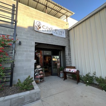 Cave & Post Trading - New Location - The photo depicts the entrance of Cave & Post Trading Co., a curated men's goods store. The storefront has a sign with the business name hanging above the door. There is a small outdoor display stand with items and a wooden table with brochures or menus. The building is constructed with concrete blocks, and there are plants on either side of the entrance. The sky is clear and blue, indicating a sunny day. - Men's Clothing Near Me - Phoenix, Arizona