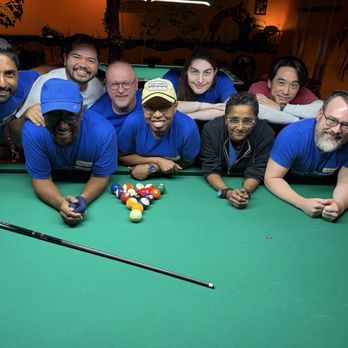 Atomic Billiards - A group of nine people, including men and women, are gathered around a pool table at Atomic Billiards in Washington, DC. They are leaning on the table, smiling at the camera, with a set of pool balls arranged in the center. Some are wearing blue shirts, and one person has a cap. The background shows a dimly lit bar environment with orange walls and a television screen on the right side. The scene captures a social moment in a billiards and bar setting. - Pool Halls Near Me - Washington, DC, District of Columbia