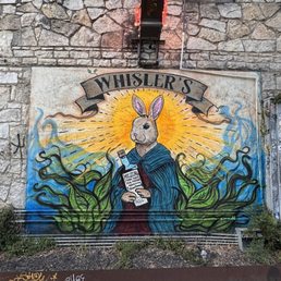 A colorful mural on a stone wall depicts a rabbit with a halo, holding a mezcal bottle, surrounded by green foliage and a bright background, with "Whisler's" written above.