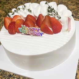 Strawberry Cream Cake - A white, round birthday cake topped with fresh strawberries, whipped cream, and small green herbs, with a colorful "Happy Birthday" sign.